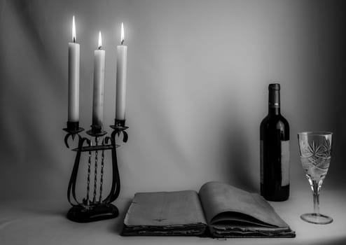 A black and white still life composition featuring an open vintage book, an antique candelabrum with burning candles, a wine bottle, and a glass, creating a timeless and moody atmosphere