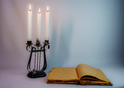 Antique candelabrum with three lit candles beside an aged open book with yellowed pages. A nostalgic, historical, and mysterious atmosphere, perfect for vintage and gothic themes