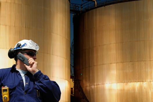 engineer, worker in hardhat, in front of sunlit large fuel-storage towers, tanks, inside oil and gas refinery
