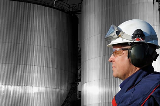 engineer, in profile, large industrial fuel-storage tanks in background, sunlight hitting the metal