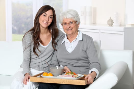 Elderly woman and home carer sitting in sofa with lunch tray