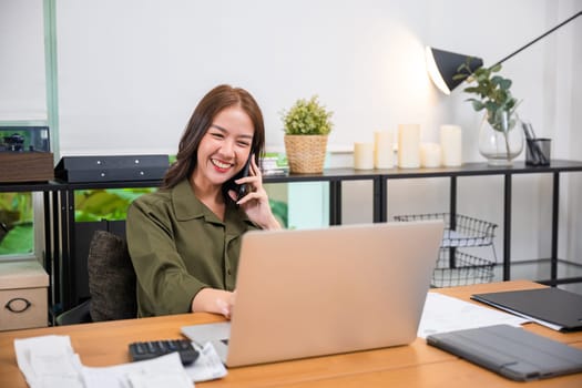 Happy Asian young woman working on laptop computer at home office while talking on mobile phone, smiling businesswoman having business call to talking with company sales client financial