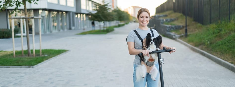 A woman rides an electric scooter with a dog in a backpack. Pappilion Spaniel Continental in a sling. Widescreen