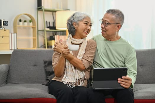 Aged spouses relaxing in living room and using digital tablet. Retirement lifestyle concept.