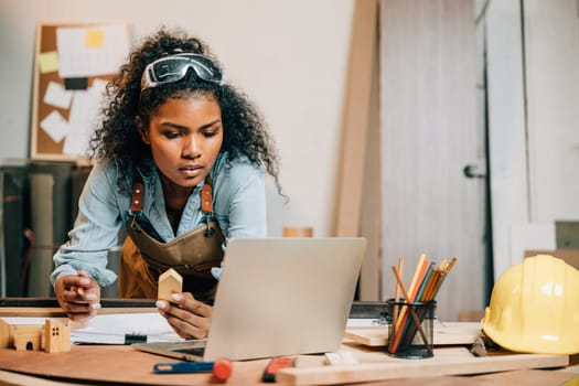 Carpenter america black woman curly hair sketch making notes in work paper while standing at wooden table with laptop computer, young female working learning online at woodshop, Happy Carpenters Day