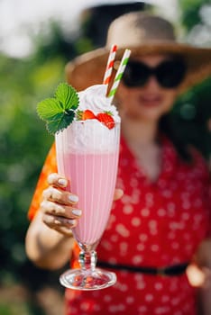 Pretty woman offers strawberry milkshake cocktail into camera, nature backdrop. High quality photo