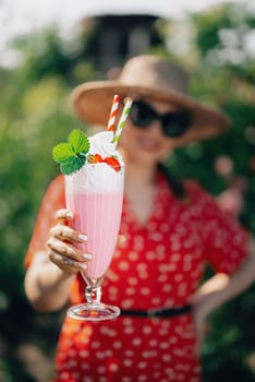 Pretty woman offers strawberry milkshake cocktail into camera, nature backdrop. High quality photo