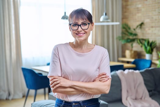 Portrait of positive confident middle aged woman at home. Smiling successful female with crossed arms looking at camera, living room interior background