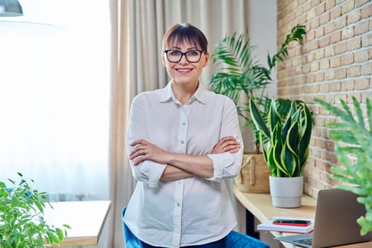 Portrait of positive confident middle aged woman at home. Smiling successful female with crossed arms looking at camera, living room interior background