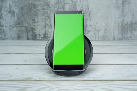 Modern smartphone with a bright green screen placed on a wireless charging pad on a wooden surface, with a textured gray background, ideal for mockups and design projects.