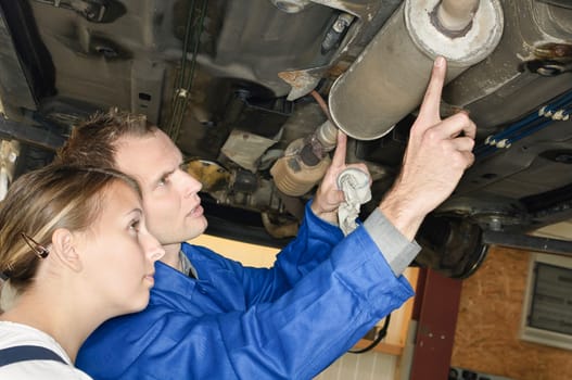 The master mechanic shows the young trainee how to test the exhaust system on the underbody in the car workshop.