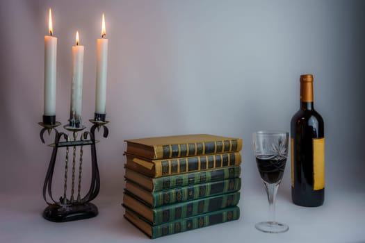 A stack of vintage hardcover books, an ornate candelabrum with burning candles, a wine bottle, and a filled glass, set in dim lighting, creating a nostalgic and elegant atmosphere
