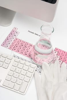 Chemist workplace - chemical flask with clear liquid, periodic table of elements, protective gloves and computer