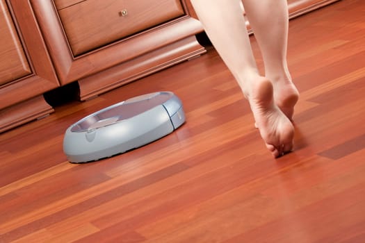 Woman go round home washing robot in cleaning action, saving her time. Genuine living room wooden floor (jatoba). Selective focus on robot.
