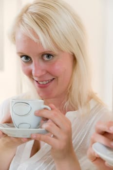 Portrait of young blonde woman holding coffee cup in front of bright kitchen in background and looking at camera