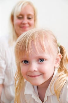 Head and shoulders portrait of a little blonde four-year-old girl in wide angle with mother in blurred background against white wall