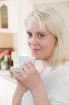 Portrait of young blonde woman holding coffee cup in front of bright kitchen in background and looking at camera