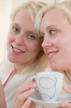 Head and shoulders portrait of a young blonde woman holding a coffee cup in front of mirror in original and mirror view looking at camera