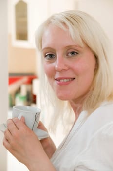 Portrait of young blonde woman holding coffee cup in front of bright kitchen in background and looking at camera