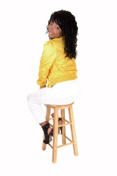 A young pretty African American woman sitting on an chair in white  tights and a yellow jacket from the back, looking over her shoulder,  for white background.
