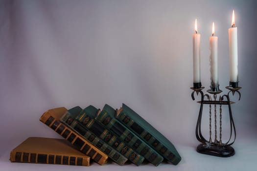 A stack of antique hardcover books tipping over, next to a vintage candelabrum with burning candles, creating a nostalgic and dramatic atmosphere