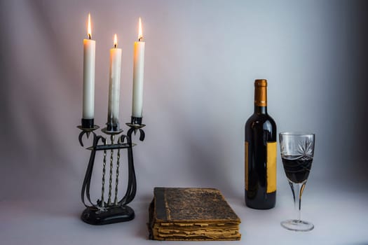 A stack of antique books, an ornate candelabrum with burning candles, a wine bottle, and a filled glass, set in dim lighting, creating a sophisticated and moody atmosphere
