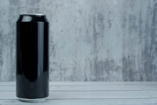 Blank black aluminum can standing on a wooden surface against a gray textured wall, suitable for branding and product mockup.