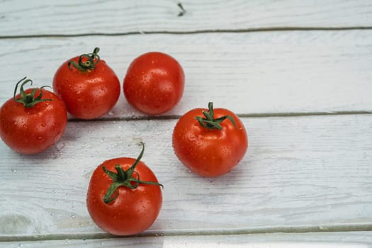 Close-up of five fresh red tomatoes with green stems and water droplets on a rustic white wooden background, creating a natural and organic look.