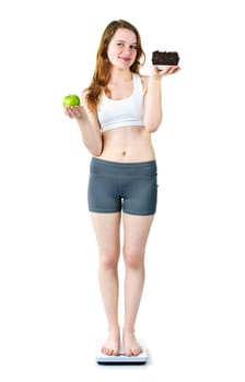 Tempted young woman holding apple and chocolate cake standing on scale