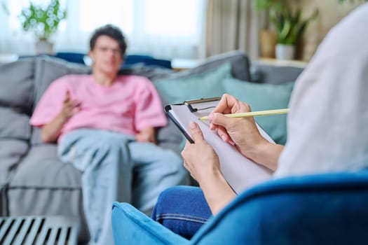 Close-up of female psychologist's hands with clipboard making notes, in individual therapy session. Young male patient in chair out of focus. Psychology psychotherapy treatment mental health concept