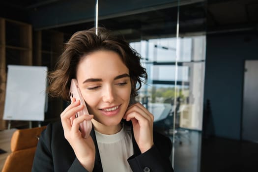 Portrait of young corporate woman, businesswoman in suit, answers phone call and smiles, stands in office conference room. Business and people concept
