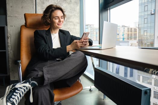 Portrait of stylish businesswoman working in office, using empty coworking room, sitting with laptop and smartphone. Corporate people concept