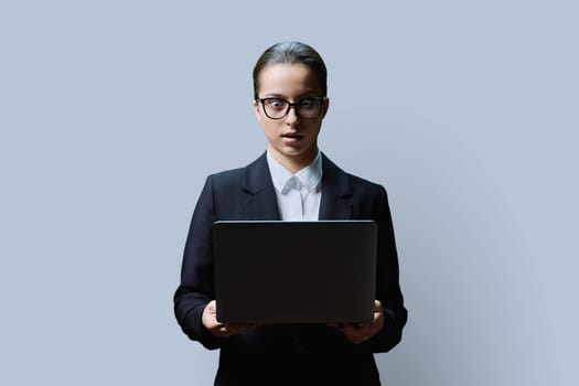 Serious teenage female student in black jacket using laptop on grey studio background. Education, learning, technology, knowledge concept