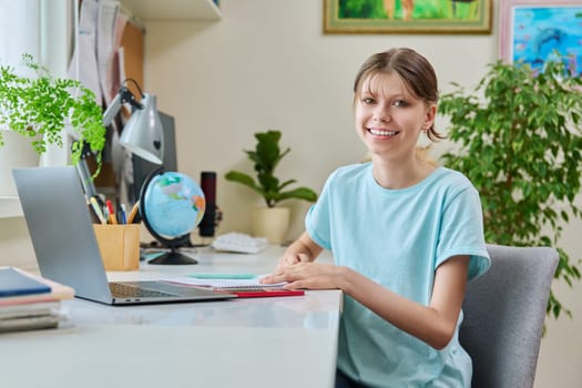 Portrait of teenage girl sitting at desk at home with laptop computer. Smiling 12, 13 year old young female student looking at camera. High school, adolescence, education, lifestyle concept