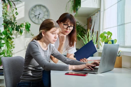 Mother and preteen daughter looking into laptop computer together, girl typing on keyboard, sitting at desk at home. Family, home lifestyle, study, kids concept