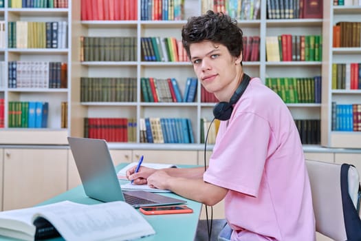 Young male student sitting with computer laptop with books in college library. Guy 18-20 years old with headphones looking at the camera. Knowledge, education, youth, college university concept