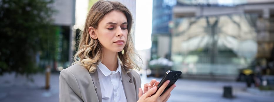 Young businesswoman looking complicated, frowning as reading message on mobile phone. Serious corporate woman checking her smartphone.