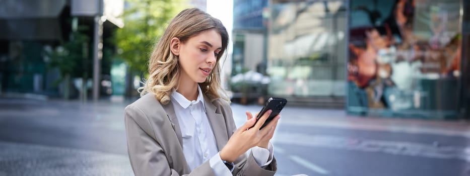 Blond woman in suit, looking at her mobile phone, order taxi, using application, posing outdoors.