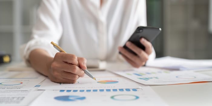 Accountant using mobile phone at office, analyzing business document with graph. Financial chart, marketing report. Accounting data analysis concept.