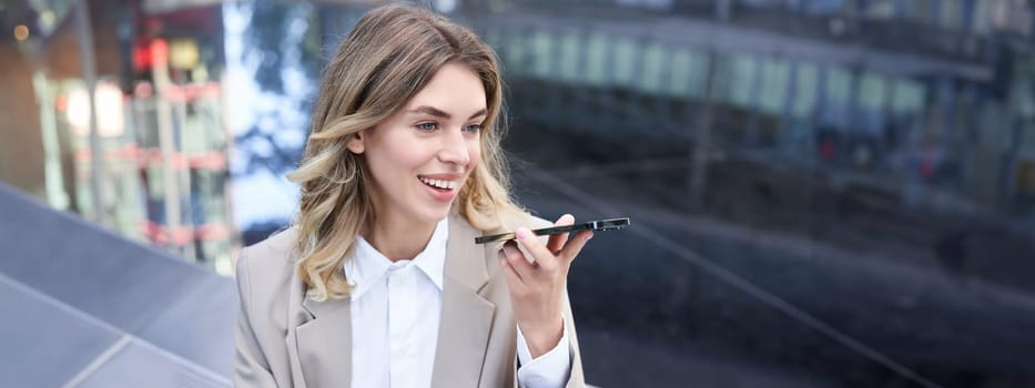 Stylish corporate woman records voice message on smartphone, talks into microphone on mobile phone, holds telephone near lips while speaking, sits alone near fountain.