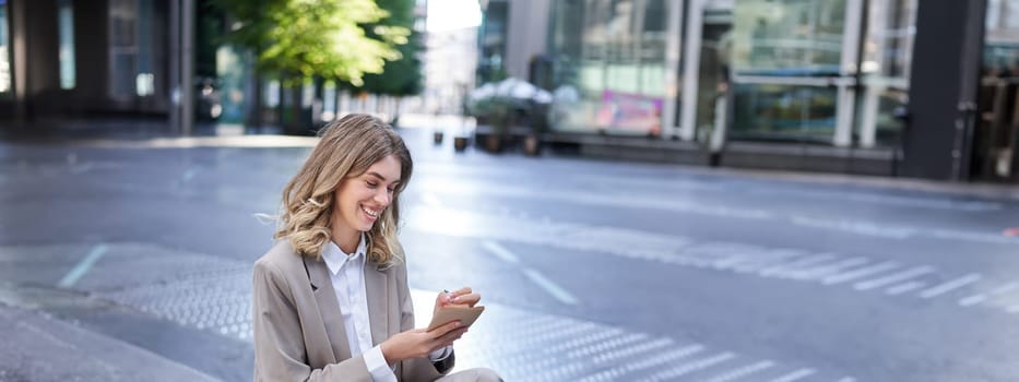 Businesswoman sitting with laptop, wearing suit, writing down notes in her planner, being in city centre outdoors.