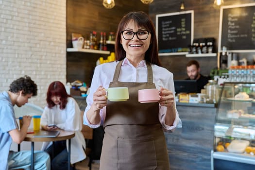 Portrait of smiling female coffee shop worker holding two cups of coffee in her hands. Woman in apron looking at camera inside cafe hall. Small business, food service occupation, staff, work concept