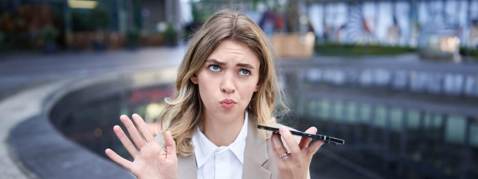 Portrait of confused businesswoman recording voice message, counting, thinking while talking into mobile phone, sitting near ofice buildings on street.