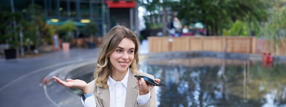 Smiling business woman record voice message, speaking into microphone on mobile phone, sitting near fountain on street.