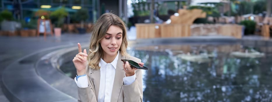 Vertical shot of businesswoman in suit, talking in microphone on phone, record voice message, using speakerphone while sitting on empty street near office buildings.