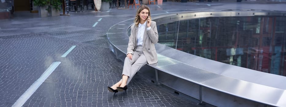 Young female manager. Corporate woman sitting outside on street and talking on mobile phone. Girl making phone call.