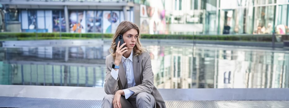 Stylish businesswoman in suit, sitting near fountain outside office buildings, talking on mobile phone. Corporate woman waiting for someone outside.