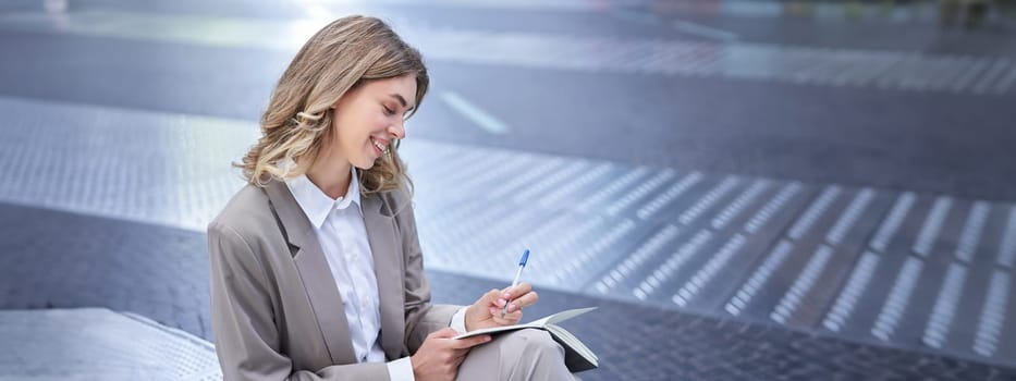 Business woman in suit sits in city centre, writes down, takes notes, holds pen and notebook, brainstorms, creates ideas.