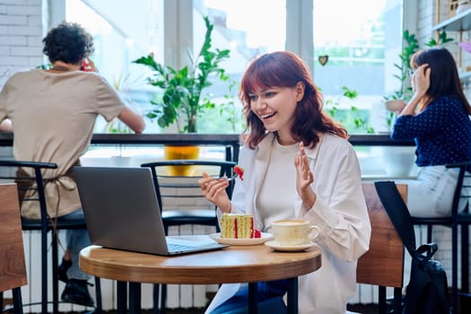 Female college student with laptop in cafe at table with cup of coffee and piece of cake. Internet online technology for leisure communication blogging learning chat, youth lifestyle concept