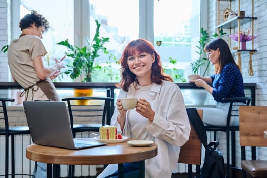 Female college student with laptop in cafe at table with cup of coffee and piece of cake. Internet online technology for leisure communication blogging learning chat, youth lifestyle concept
